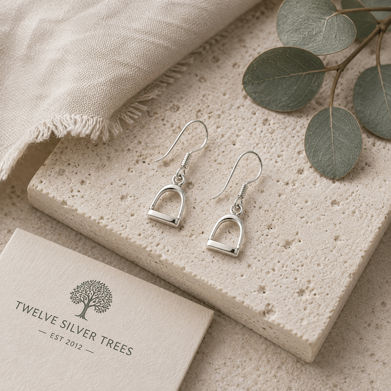 Small sterling silver stirrup hook earrings on soft stone flatlay styled with linen and eucalyptus by Twelve Silver Trees