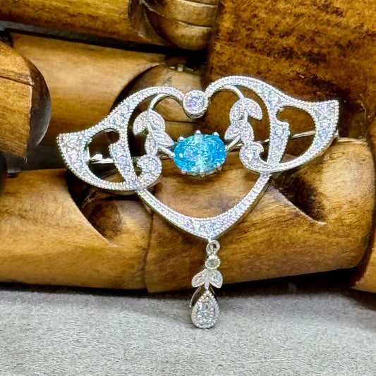  a belle epoque style art nouveau brooch, handcrafted in sterling silver and set with fine zirconia. The brooch features clear gemstones on the garlands and swags with a central old cut oval blue gemstone