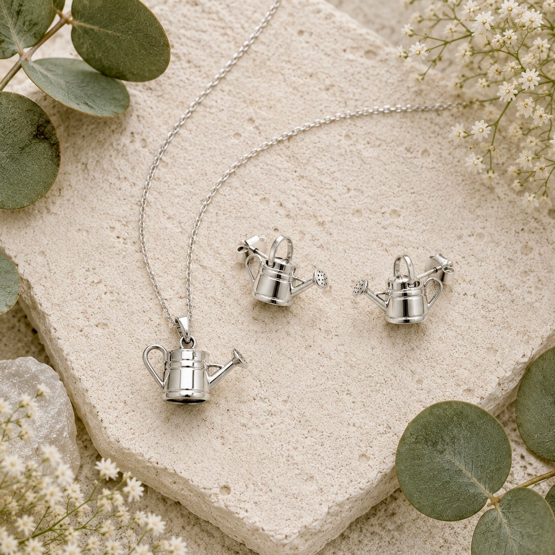 Sterling silver watering can pendant necklace and matching stud earrings styled on natural stone with eucalyptus and white flowers.